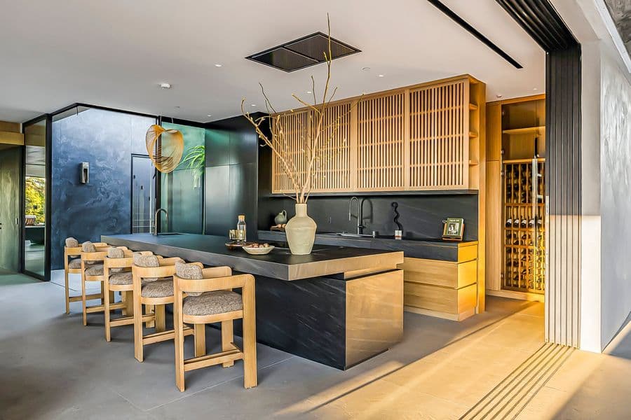 Bar with slatted oak upper cabinets, a black stone island and counters, oak stools and a lit, floor-to-ceiling wine wall