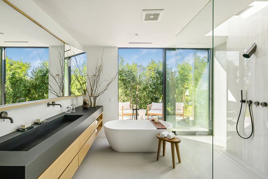 Bathroom with a long wall-hung oak vanity under a black trough sink, a wide mirror, and a freestanding tub by a window