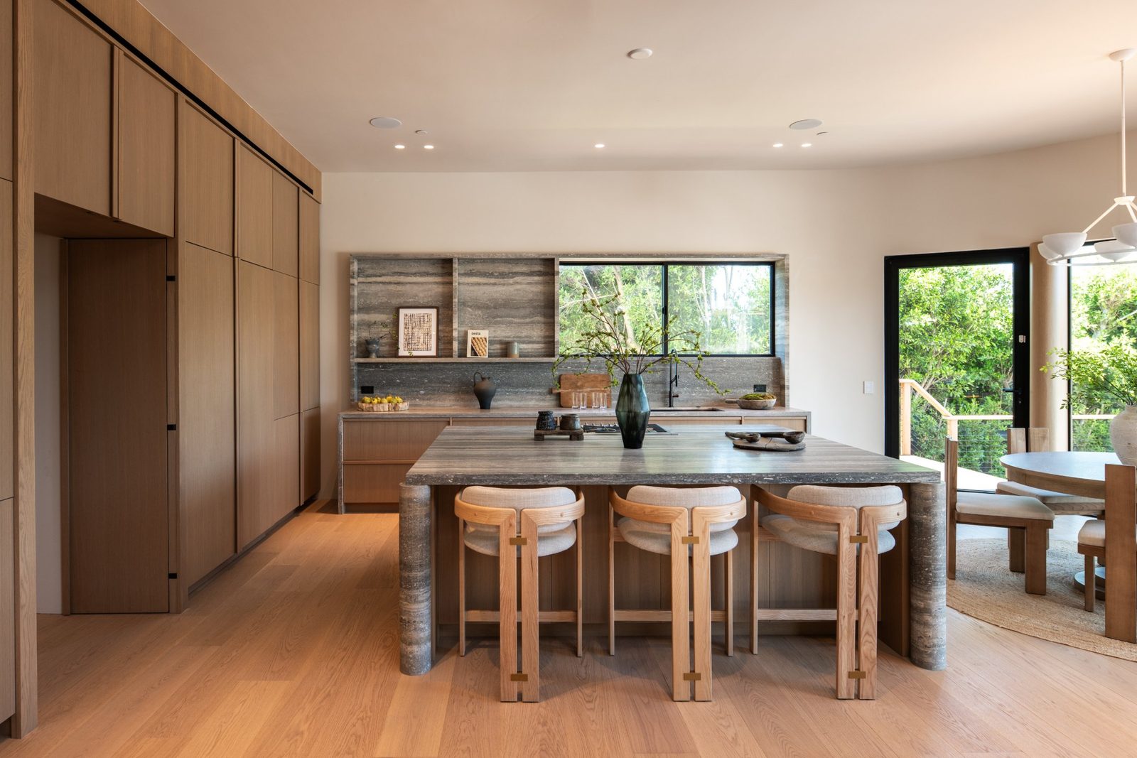 Kitchen with a full-height wall of rift white oak cabinets, a travertine island with upholstered stools, and a travertine backsplash under a wide window